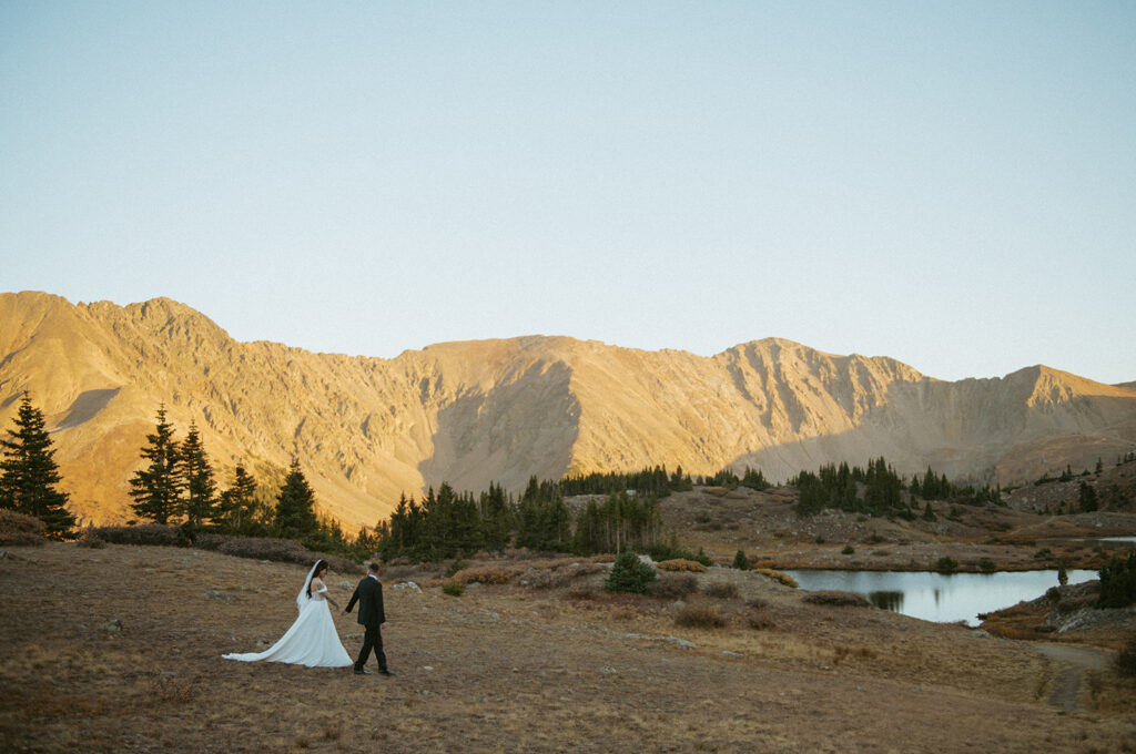 A couple walking hand in hand on their Colorado elopement day near a lake with mountains during sunset