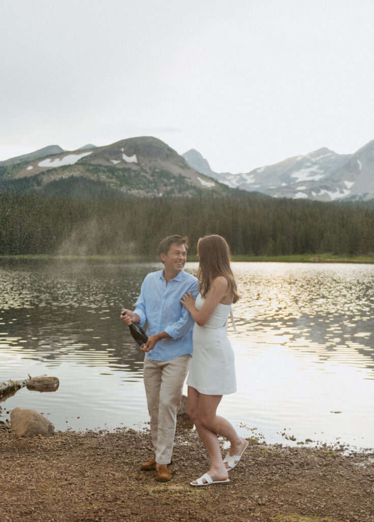 A couple pops a champagne bottle along the shoreline to celebrate their engagement session