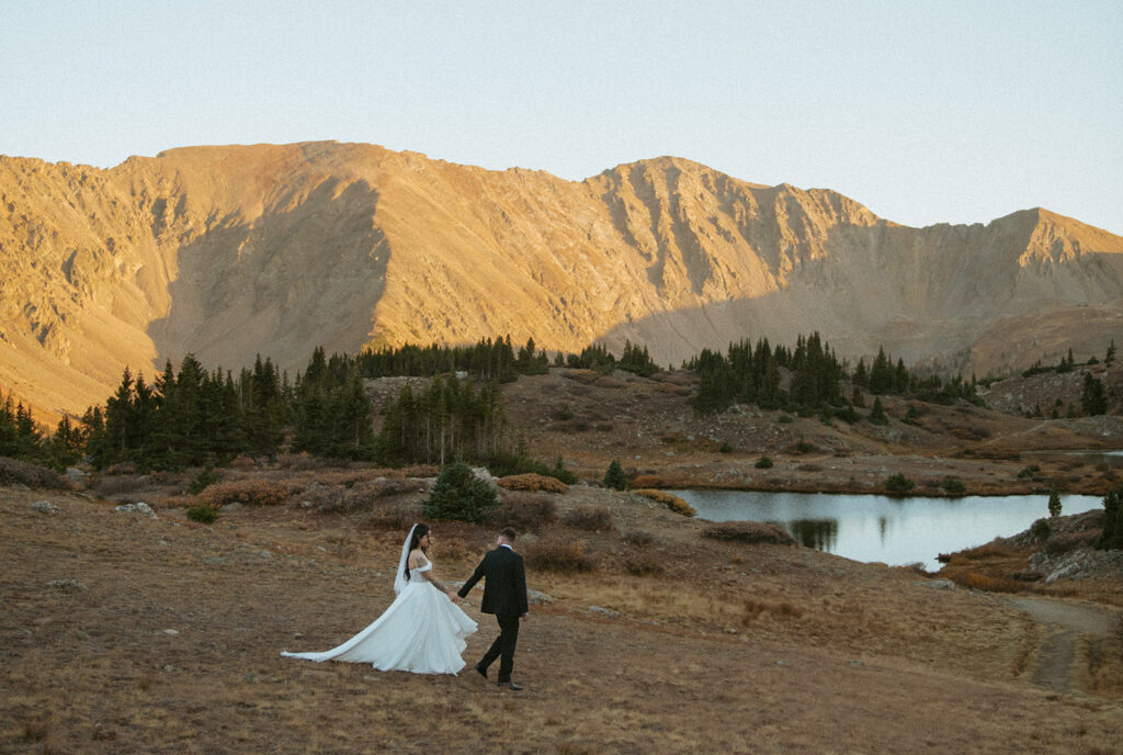 A couple hand in hand on their Colorado elopement day, surrounded by an alpine lake and mountains at sunset