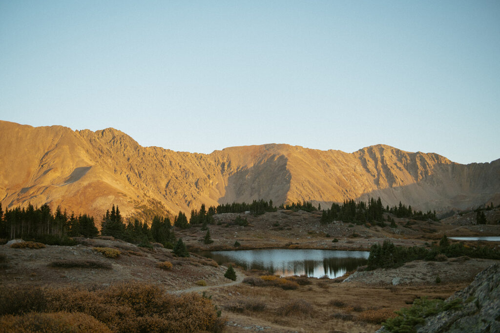 The sunset paints the mountains an orange hue behind an alpine lake at this Colorado elopement location