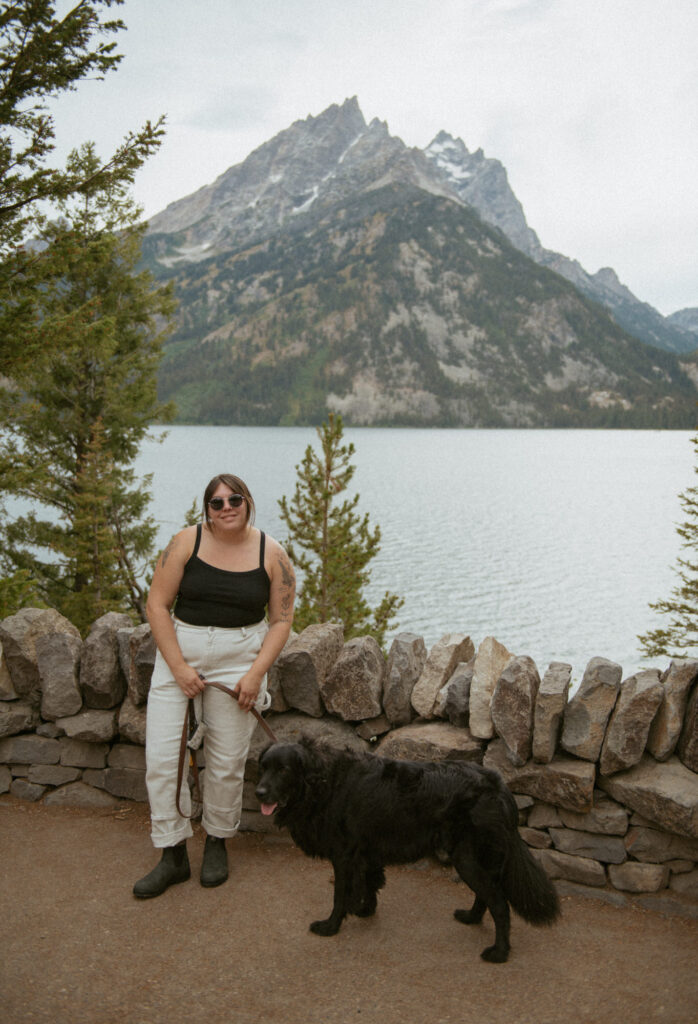 A portrait of Rachel, Colorado elopement photographer, and her dog standing by an alpine lake with a dramatic mountain view